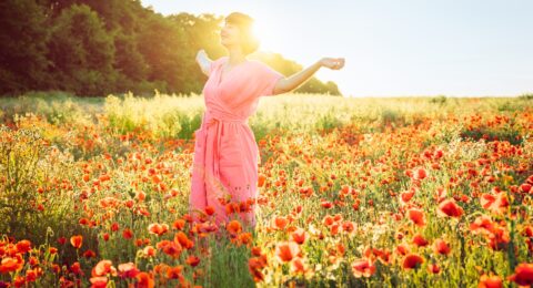 A happy young woman in a pink dress with raised arms relaxing in red poppies flowers meadow in sunset light. A simple pleasure for mental health. Nature relaxation. Selective focus. Copy space.