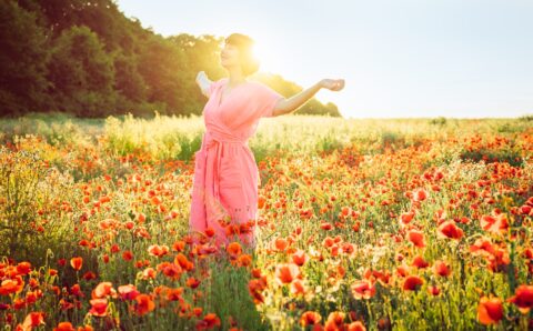 A happy young woman in a pink dress with raised arms relaxing in red poppies flowers meadow in sunset light. A simple pleasure for mental health. Nature relaxation. Selective focus. Copy space.
