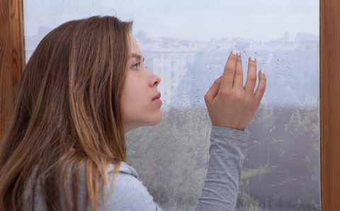 Lonely young lady looking out window, lost in thought, touching glass with rain drops