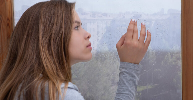 Lonely young lady looking out window, lost in thought, touching glass with rain drops