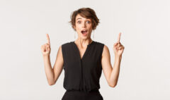 Portrait of excited and amused young woman gasping fascinated while pointing up, standing white background