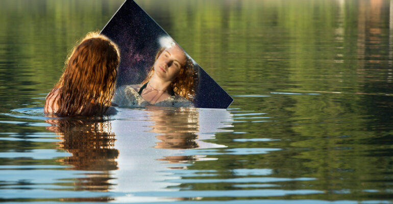 Young woman standing in lake, holding mirror, looking at reflection